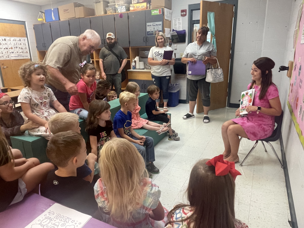 Teacher in pink dress holding book in front of new kindergarten class